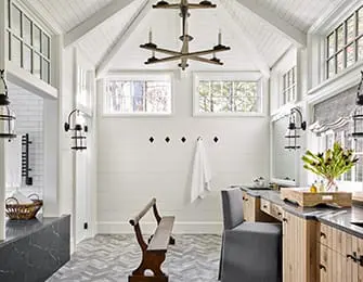 Bright white room with shiplap walls, vaulted ceiling, and herringbone tile floor. Features a wooden bench, a hanging towel, and a vanity area with a stone countertop.