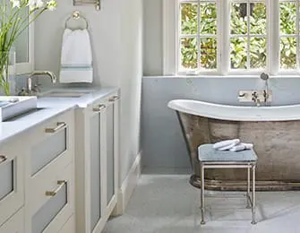 Light blue bathroom with a freestanding tub, white vanity with silver hardware, and a window overlooking greenery.