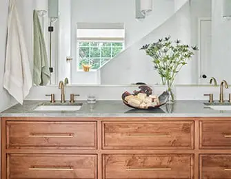 Double vanity bathroom with warm wood cabinetry, brass hardware, and gray stone countertops. A vase of greenery sits on the counter, with a window reflected in the mirror.