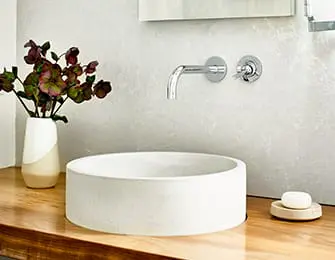 Concrete vessel sink on a wooden vanity countertop with a chrome faucet and a vase of dark purple flowers.