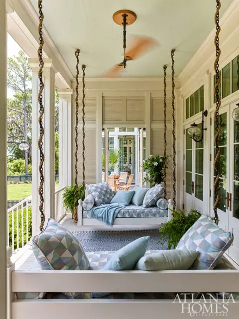 Porch swing with blue and white geometric patterned cushions and pillows, a light blue throw blanket, and a patterned rug. A ceiling fan is visible overhead.