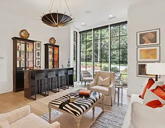 Living room with a built-in bar, zebra print ottoman, upholstered chairs, and large windows overlooking a wooded area.
