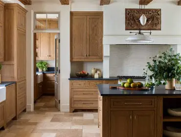 Warm wood cabinetry, a white farmhouse sink, dark countertops, and a large island define this spacious kitchen. Exposed beams and stone tile flooring add rustic charm.