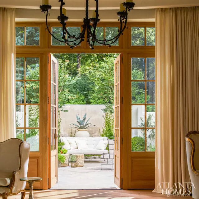View through open double doors to a patio with white sofa and potted agave plant, framed by cream curtains and a dark metal chandelier.