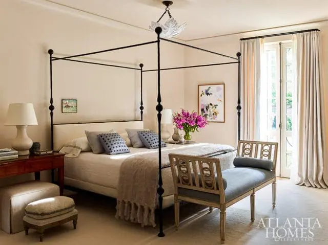 Bedroom with a black canopy bed, cream bedding, patterned pillows, and a fringed throw. A wooden bench with blue upholstery sits at the foot of the bed.
