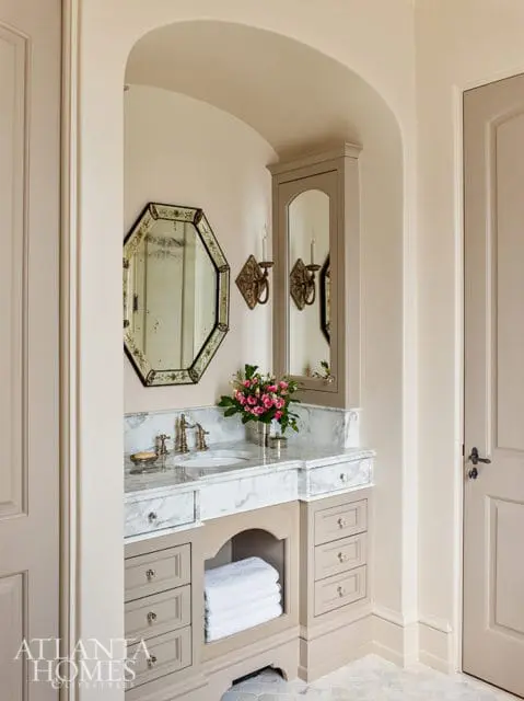 Bathroom vanity with marble countertop, brass fixtures, and taupe cabinetry. An octagonal mirror hangs above the sink, with a tall mirrored cabinet to the right.