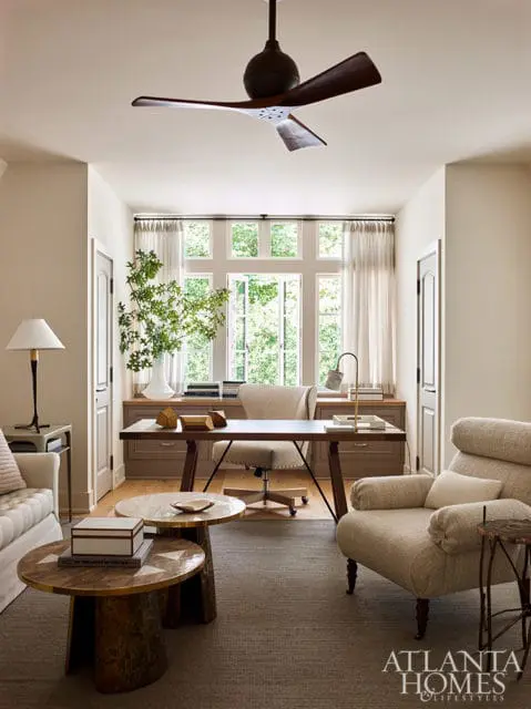 Home office with a large desk and chair facing a wall of windows, flanked by built-in cabinetry. A sofa and armchair are visible in the foreground.