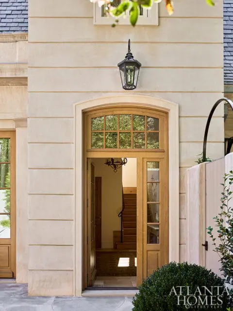 Light beige stone facade with horizontal banding, a dark lantern, and an open arched wooden door revealing a staircase.