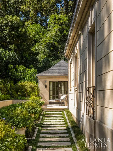 Stone pathway leading to a covered patio with a glass door and seating, flanked by lush greenery and a light-colored stone building.