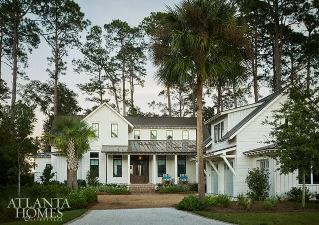 Modern farmhouse with white siding, dark metal roof, and a weathered metal porch roof. Surrounded by tall pine trees and palm trees.