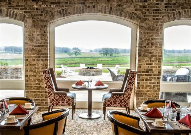 Restaurant dining room with brick walls, patterned armchairs, and tables set for a meal, overlooking a landscape with a fire pit.