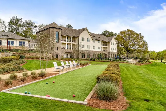 Bocce ball court with red and green balls on artificial turf, lined by hedges and chairs, with a multi-story brick and siding building in the background.