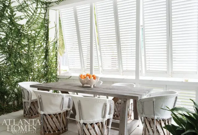 Covered porch dining area with a rustic wood table, white upholstered chairs with wood slat backs, and white louvered shutters.