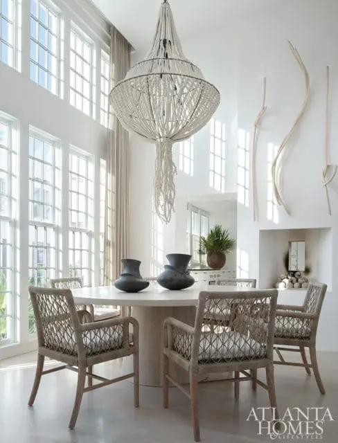 Dining room with a large white beaded chandelier, round pedestal table with two dark vases, and woven armchairs with white cushions. Tall windows and a fireplace with stacked logs are visible.