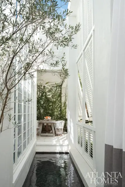 Narrow walkway with a dark water feature on the left and white shutters on the right, leading to a small patio with a wooden table, chairs, and a wall of green ivy.