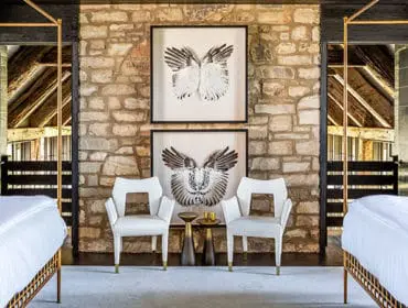 Bedroom with two gold-framed four-poster beds, white upholstered chairs, and stone accent wall featuring framed wing art.