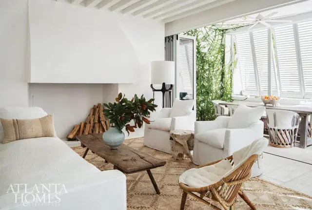 Living room with white slipcovered furniture, a large white fireplace surround, a rustic wood coffee table with a vase of greenery, and a woven rug.