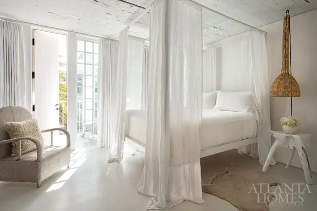 White bedroom with a four-poster bed draped in sheer white curtains, a textured armchair, and a woven pendant light.