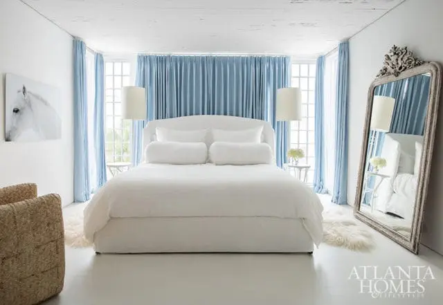 White bedroom with a large white bed, light blue curtains, a woven armchair, and a decorative mirror.