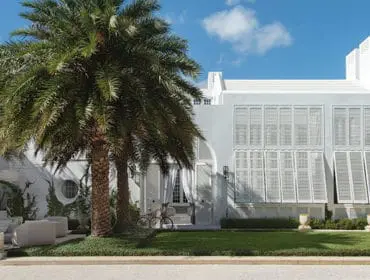 Modern white house with large louvered shutters, palm trees, and a manicured lawn.