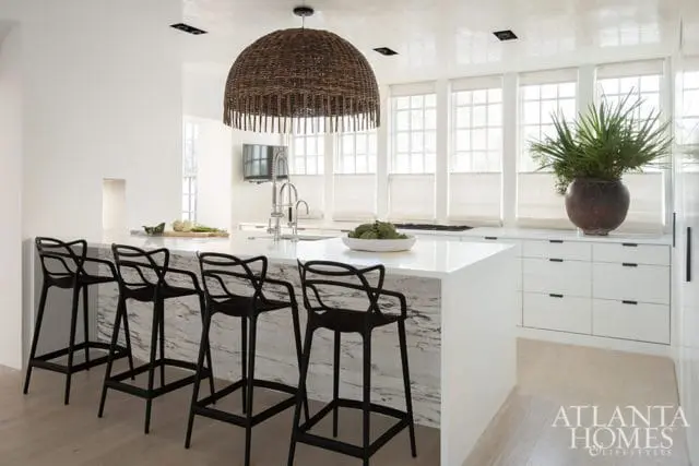 Modern white kitchen with a marble waterfall island, black bar stools, a large woven pendant light, and white cabinetry with black hardware.