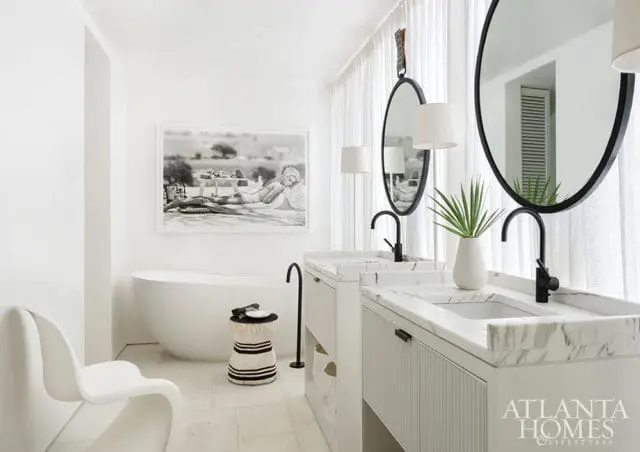 White master bathroom with freestanding tub, dual marble vanities with black faucets, oval mirrors, and a black and white framed photograph above the tub.