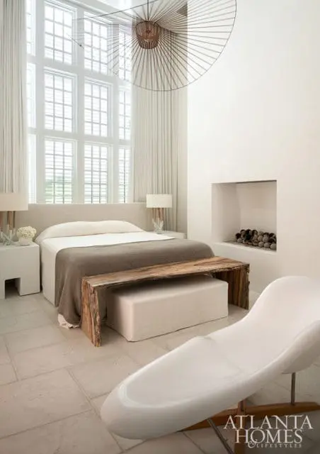 Master bedroom with a large window, upholstered bed, rustic wooden bench, and a modern white chaise lounge.