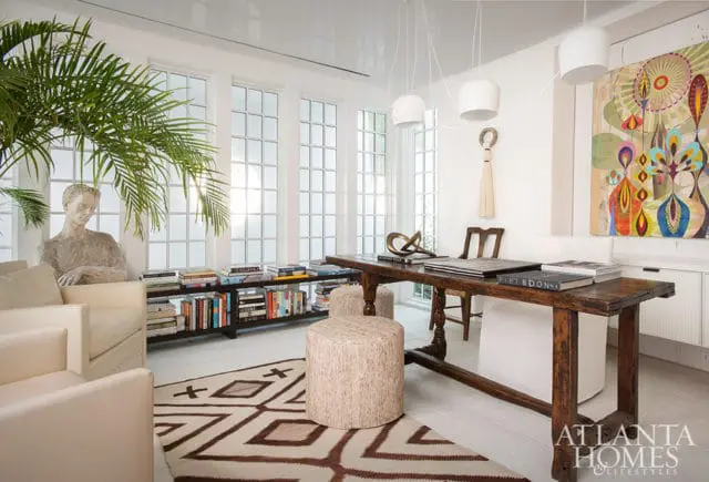 Bright office with cream armchairs, a large wooden desk, and a bookshelf filled with books. Tall windows with grid panes let in natural light, and a colorful abstract painting hangs on the wall.