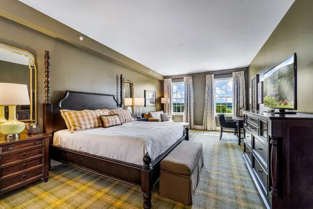King guestroom with dark wood four-poster bed, plaid pillows, green and yellow plaid carpet, and a large dresser with a television displaying a landscape scene.
