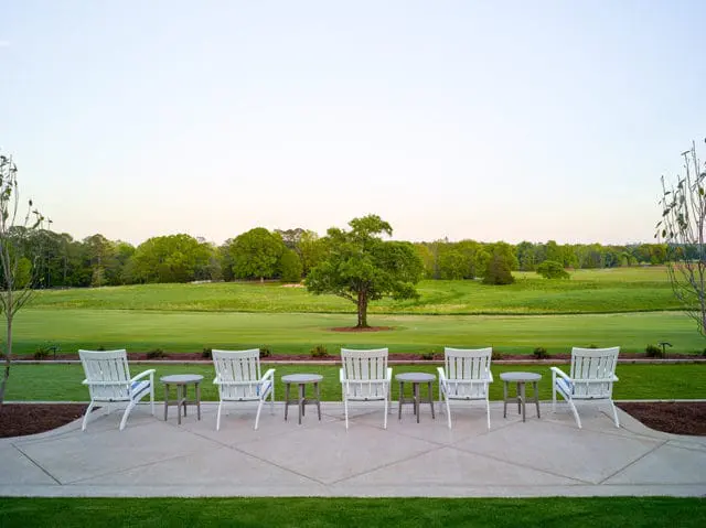White Adirondack chairs and small gray tables arranged on a concrete patio overlooking a green lawn and distant trees under a pale sky.