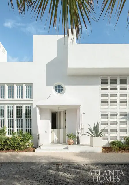 Modern white stucco house with large grid windows, louvered shutters, a round transom window, and a striped door. Lush green plants and a large agave in a planter adorn the entrance.