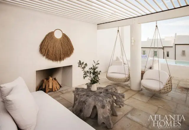Outdoor living space with white sofas, hanging rattan chairs, a stone fireplace with stacked logs, a driftwood coffee table, and a pergola overhead.