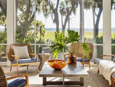 Sunroom with wicker chairs and blue cushions, a stone coffee table with a wooden bowl of fruit, and a gray sofa with a cream knit throw, overlooking a marshy landscape with trees.