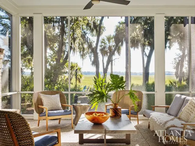 Screened porch with wicker chairs, a gray sofa with a knit throw, and a stone coffee table overlooking a marsh landscape with live oaks and palm trees.