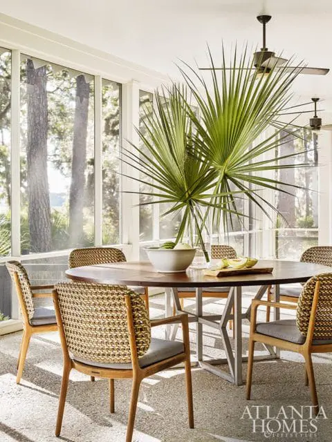 Sunroom with round wood dining table, woven chairs with gray cushions, and large palm fronds in a vase. Atlanta Homes & Lifestyles logo in the corner.