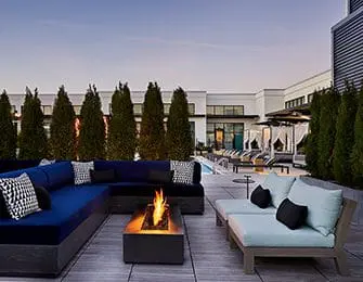Rooftop patio with a sectional sofa, lounge chairs, a fire pit, and a pool area with cabanas in the background.