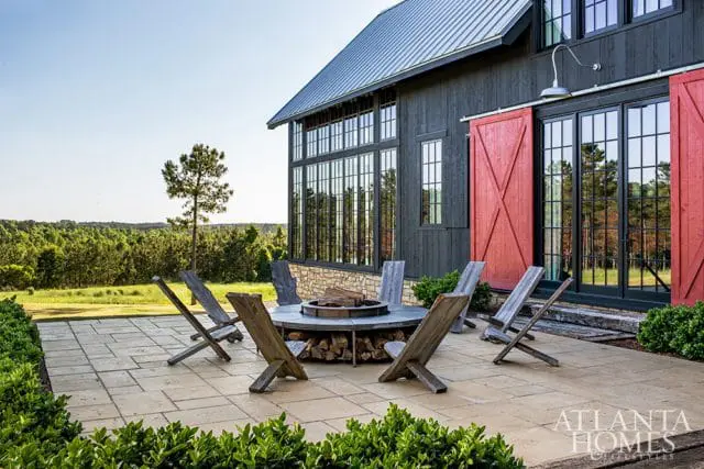 Modern farmhouse exterior with dark wood siding, large grid windows, red barn doors, and a stone base, featuring a stone patio with a circular fire pit and Adirondack chairs.