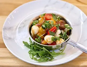 Caprese salad with cherry tomatoes, mozzarella balls, and fresh basil in a small metal bowl, served on a white plate with arugula.