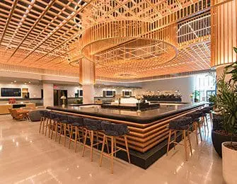 Modern bar area with a large, multi-level wood slatted ceiling, a long bar with a dark countertop, and upholstered bar stools.