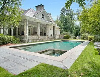 White house with stone accents and a covered porch overlooking a rectangular swimming pool surrounded by a stone patio and green lawn.