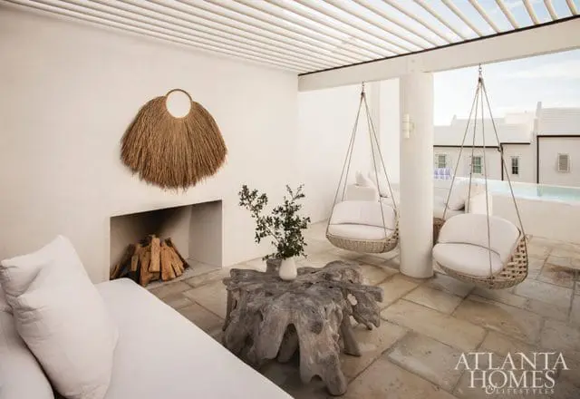 Outdoor living space with white sectional sofa, fireplace, natural wood coffee table, and two hanging rattan chairs under a slatted pergola.