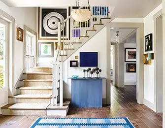 Foyer with a staircase featuring a sisal runner, abstract art, and a blue console table with decorative spheres. A blue and white patterned rug lies on the hardwood floor.