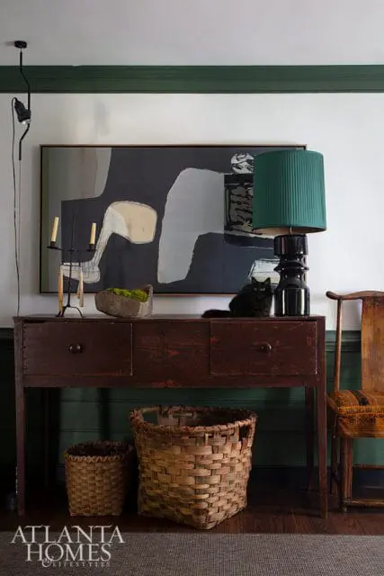 Living room with dark wood console table, abstract art, green lamp, and woven baskets. A black cat sits on the table.