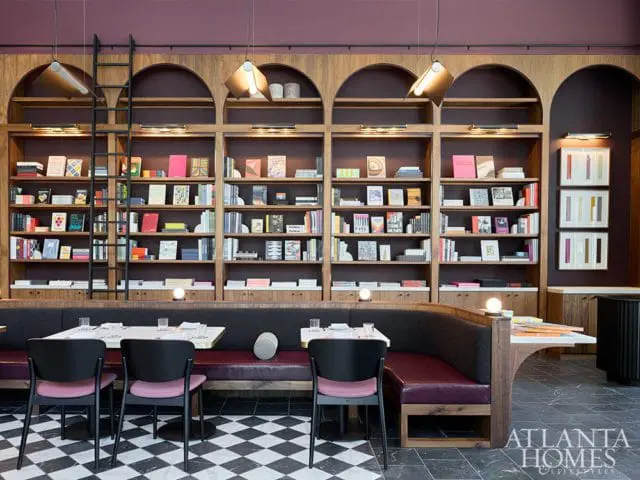 Restaurant interior with a large wooden bookshelf filled with books, a curved banquette seating area with purple upholstery, and black chairs around white tables.