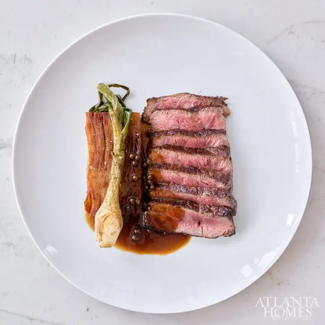 Sliced steak with sauce, roasted leeks, and peppercorns on a white plate with Atlanta Homes & Lifestyles logo in the corner