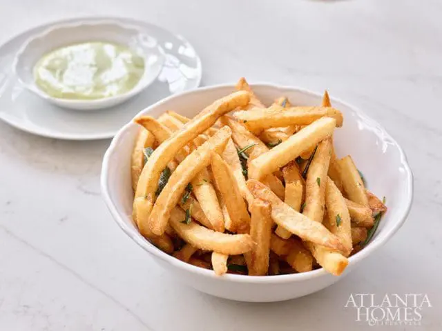 Bowl of golden french fries with herbs, next to a small dish of green sauce on a white marble surface.