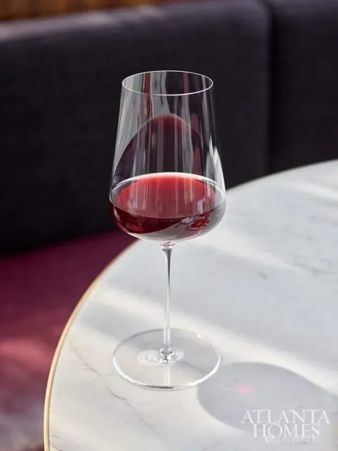 Close-up of a clear wine glass filled with red wine, resting on a white marble tabletop. The Atlanta Homes & Lifestyles logo is visible in the lower right corner.