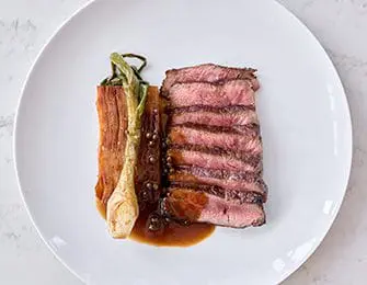 Sliced grilled steak with a side of grilled green onions and peppercorns on a white plate with a light gray background.