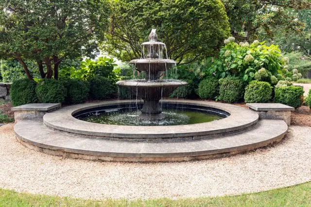 Tiered stone fountain with water cascading into a circular basin, surrounded by manicured hedges, lush trees, and hydrangeas.