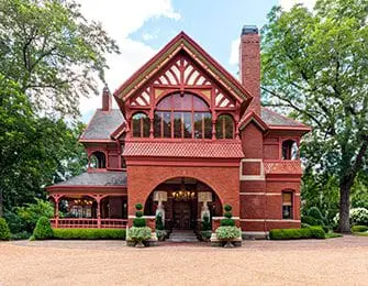 Victorian-style brick house with a red tiled roof, arched entryway, and decorative gables, surrounded by lush green trees and manicured landscaping.